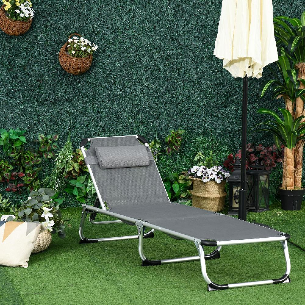 Sdraio grigio su erba sintetica davanti a siepe verde, con ombrellone e piante.