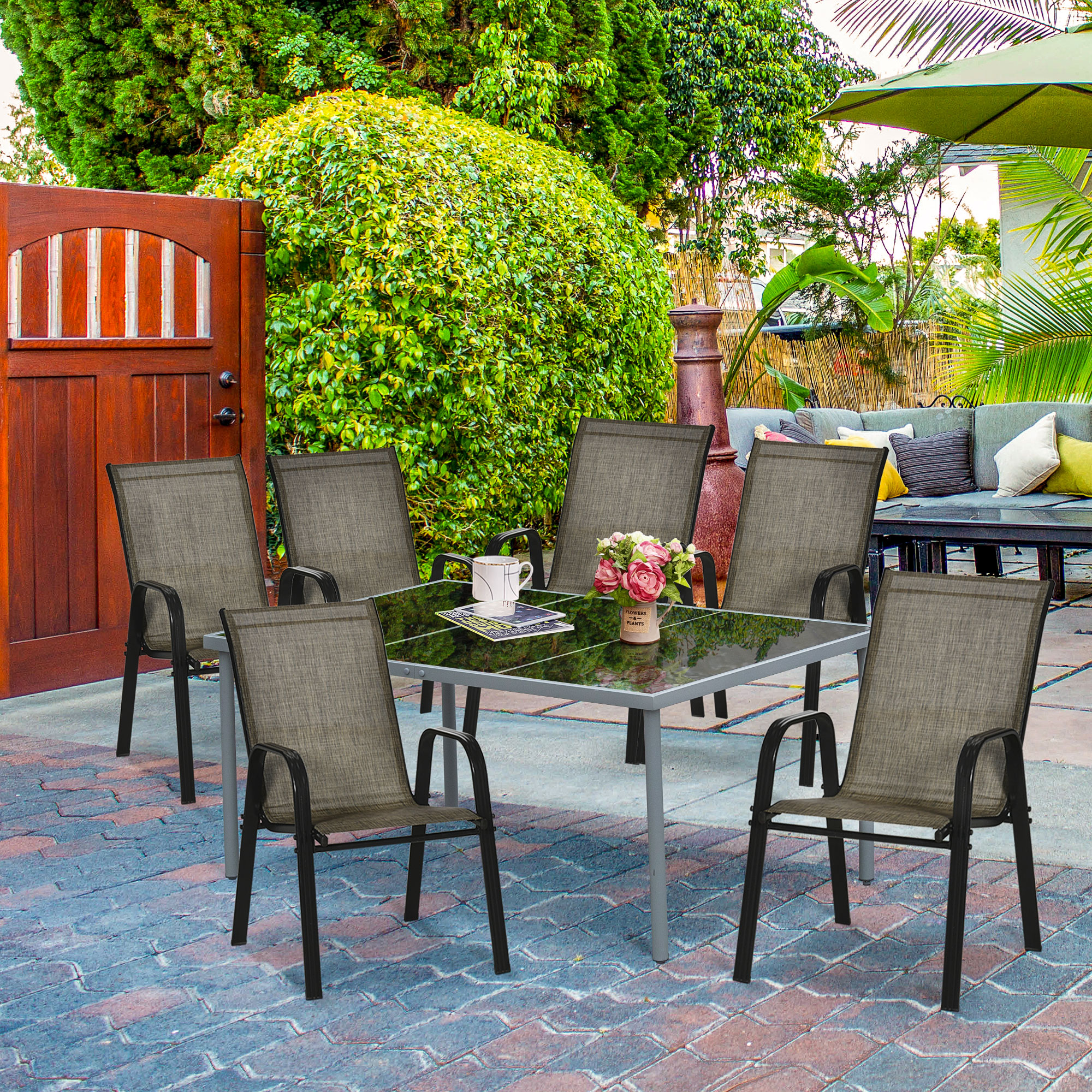 Set da patio con tavolo in vetro e sei sedie, circondato da vegetazione e cancello in legno.