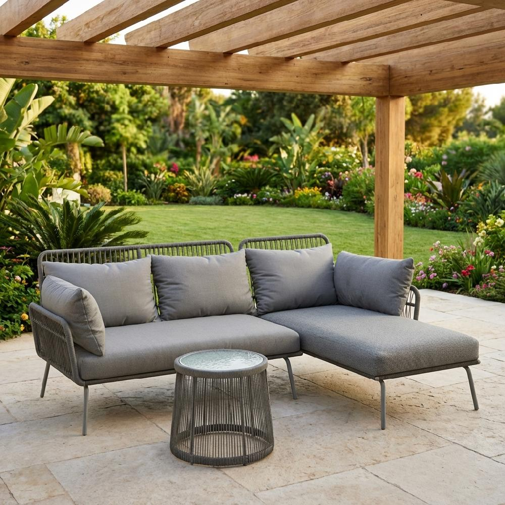 Divano da esterno angolare grigio e tavolino su patio in pietra, sfondo giardino.