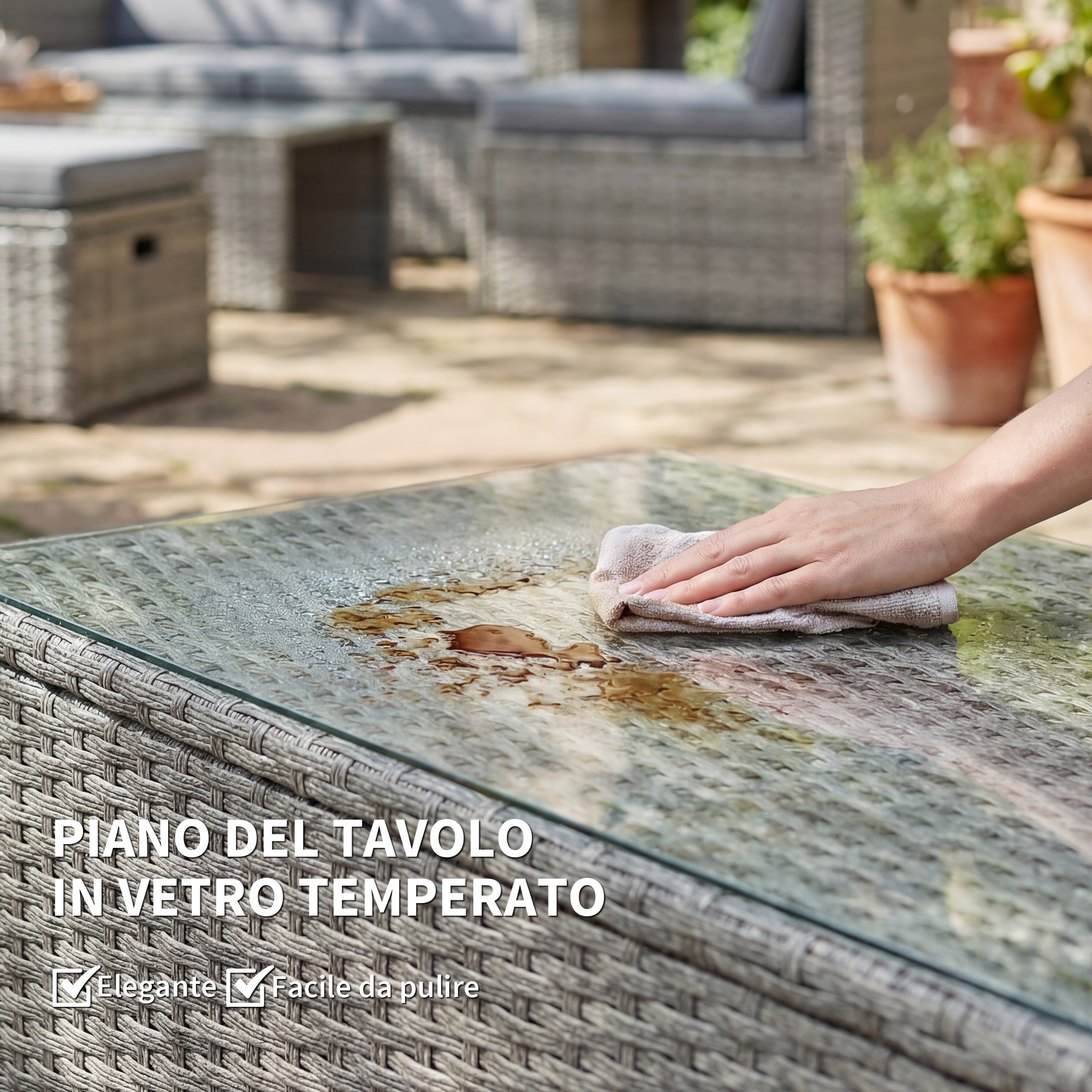 Una mano pulisce un tavolo di vetro bagnato con un panno, parte di un set di mobili in rattan.