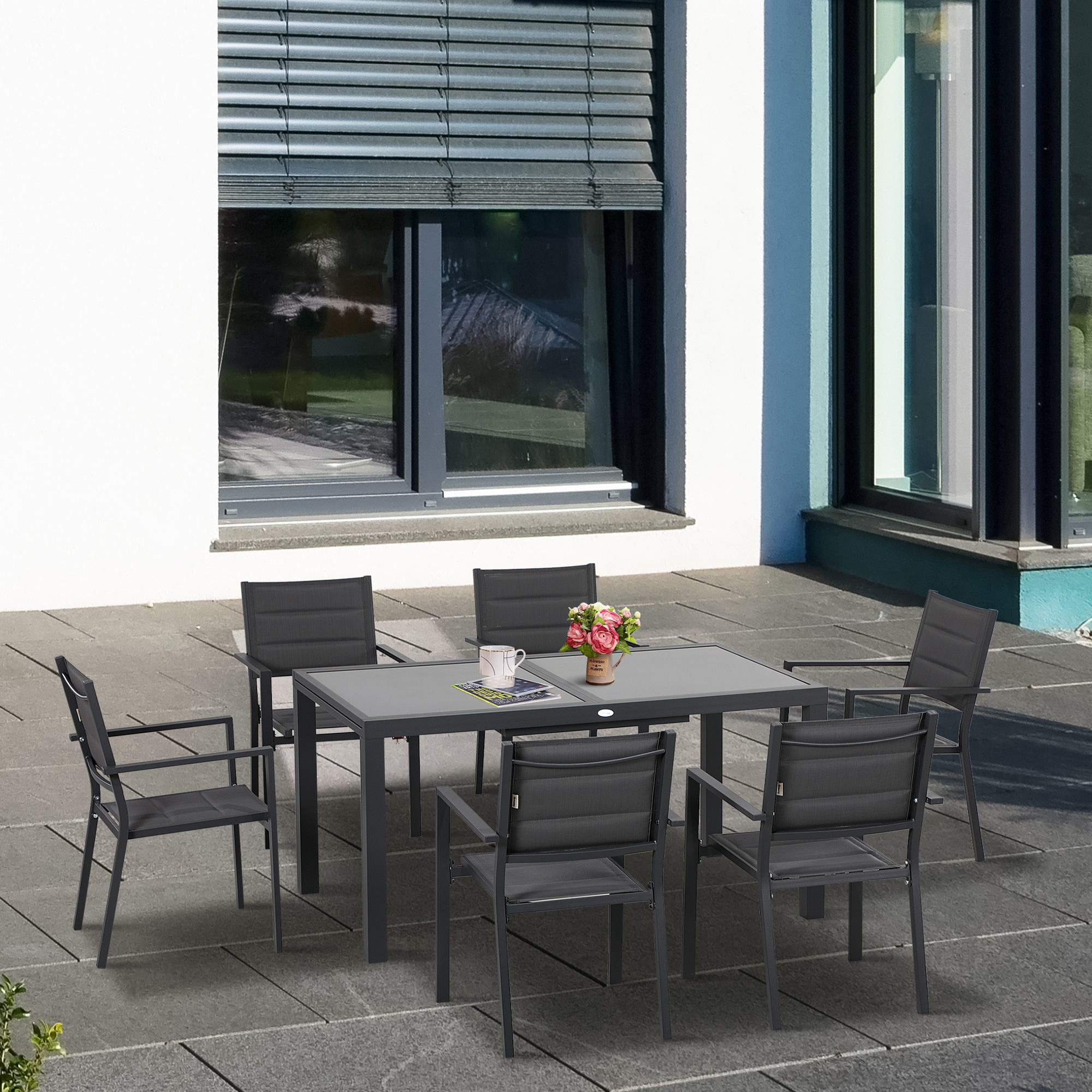 Un set da pranzo da esterno grigio con sei sedie si trova su un patio in pietra di fronte a una casa moderna.