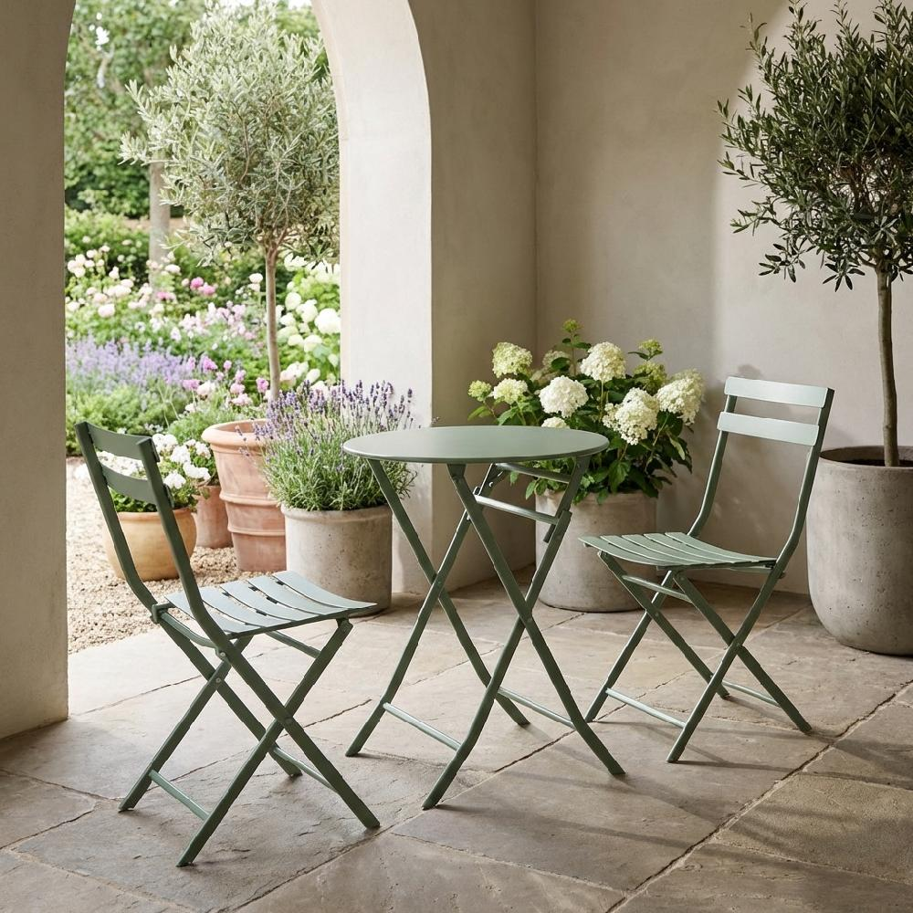 Set bistrot in metallo verde con sedie e tavolo su patio, circondato da piante in vaso e fiori.