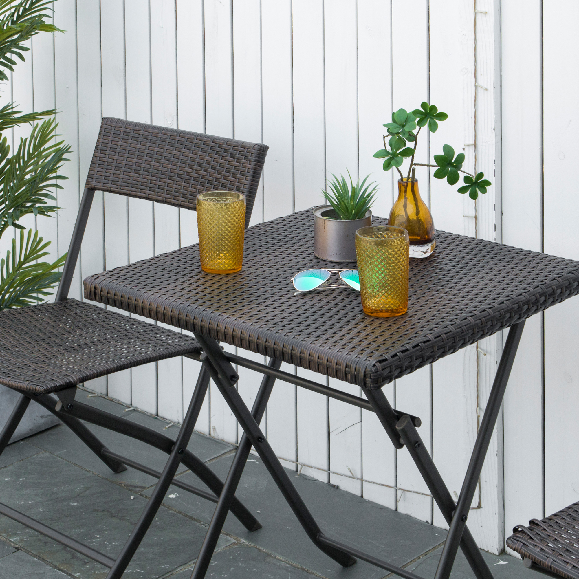 Set da patio in rattan marrone scuro con due bicchieri, occhiali da sole e piante.
