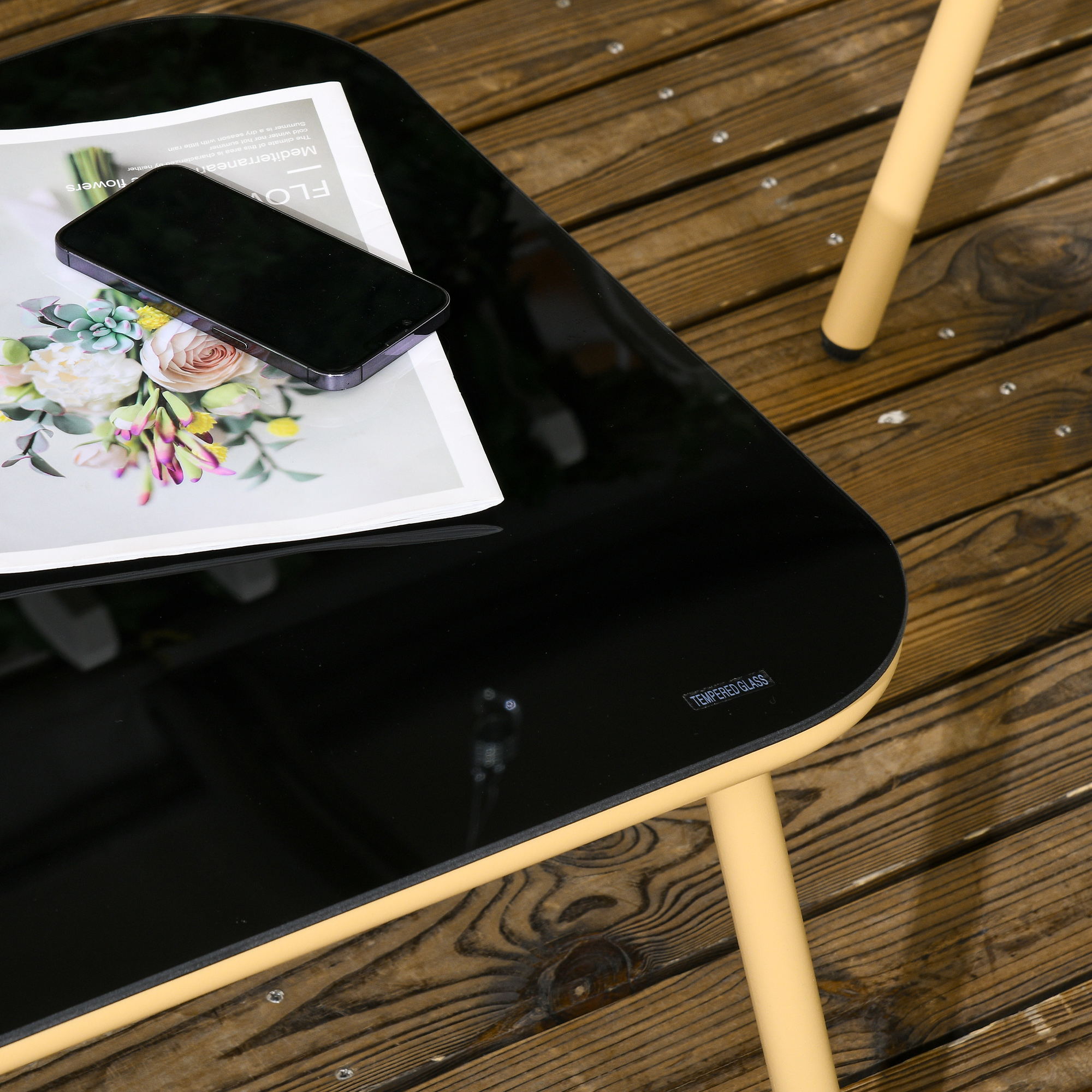 Un tavolo in vetro nero con telefono, fiori e rivista su legno.