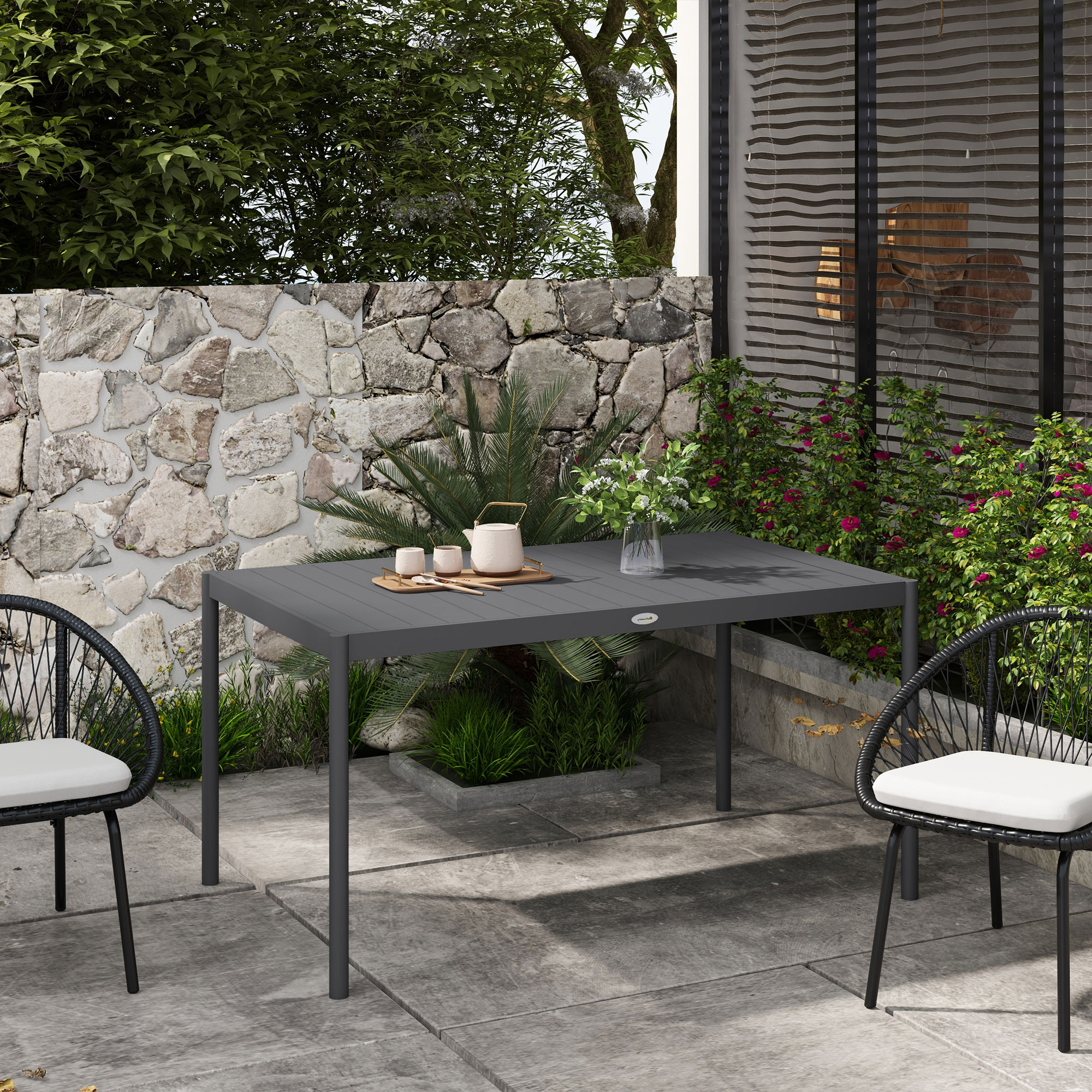 Un tavolo grigio scuro e due sedie su un patio. Un muro di pietra e piante sullo sfondo.