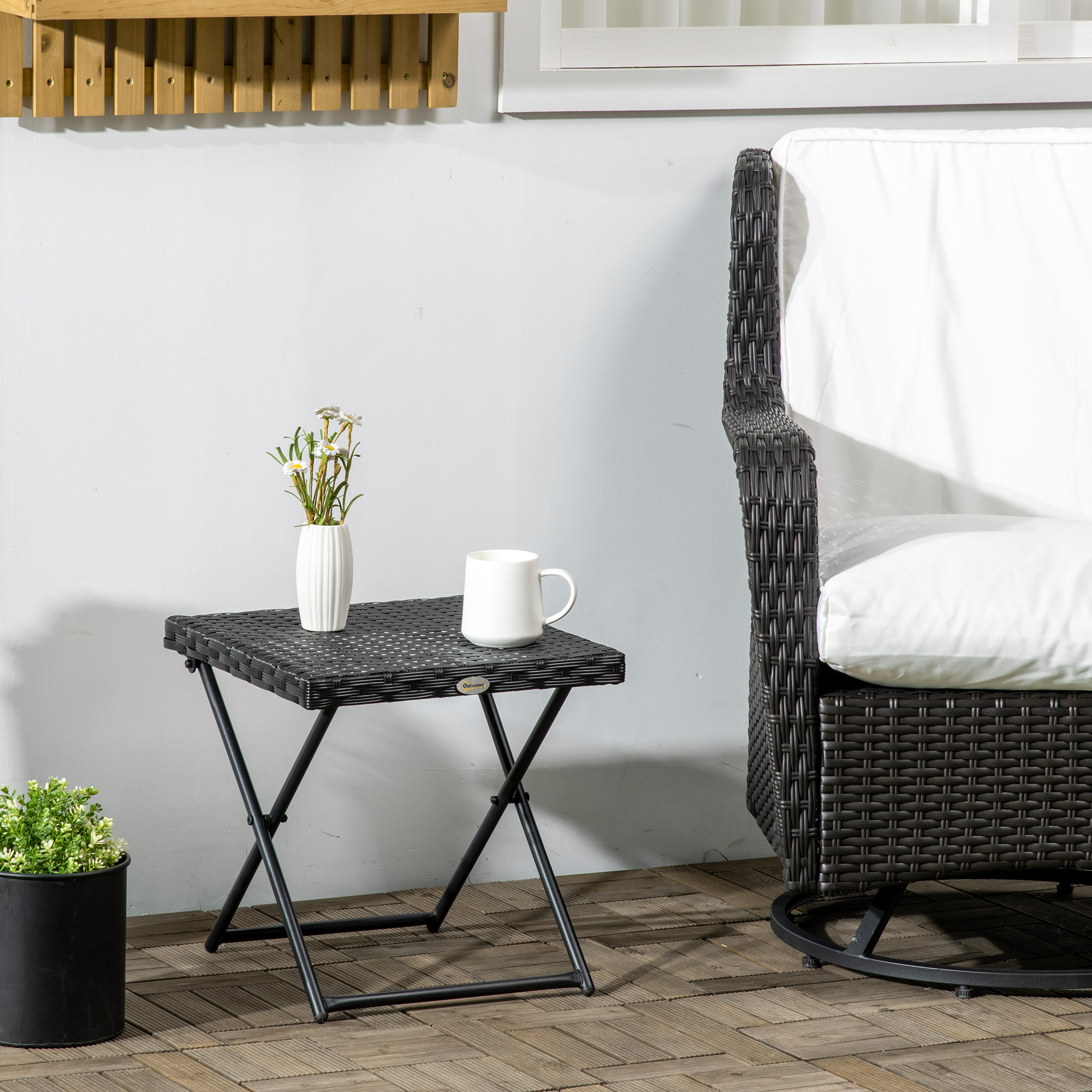 Un tavolino nero in vimini con vaso bianco, fiori e tazza. Sedia e pianta vicine.