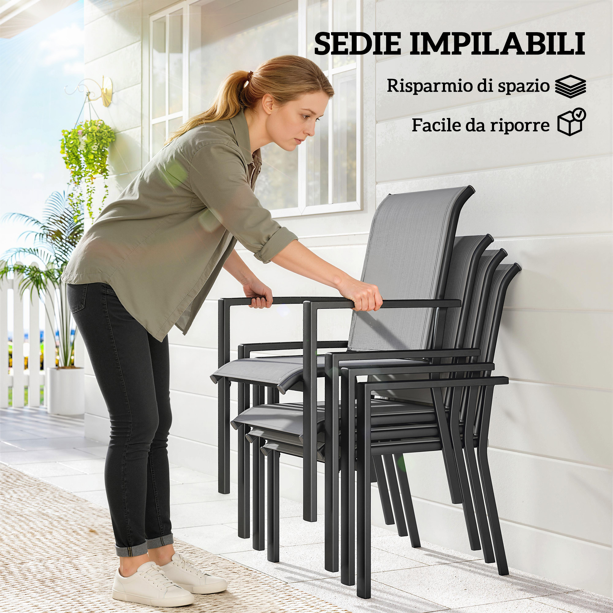 Donna impila sedie grigie da esterno su un patio con piante sullo sfondo.