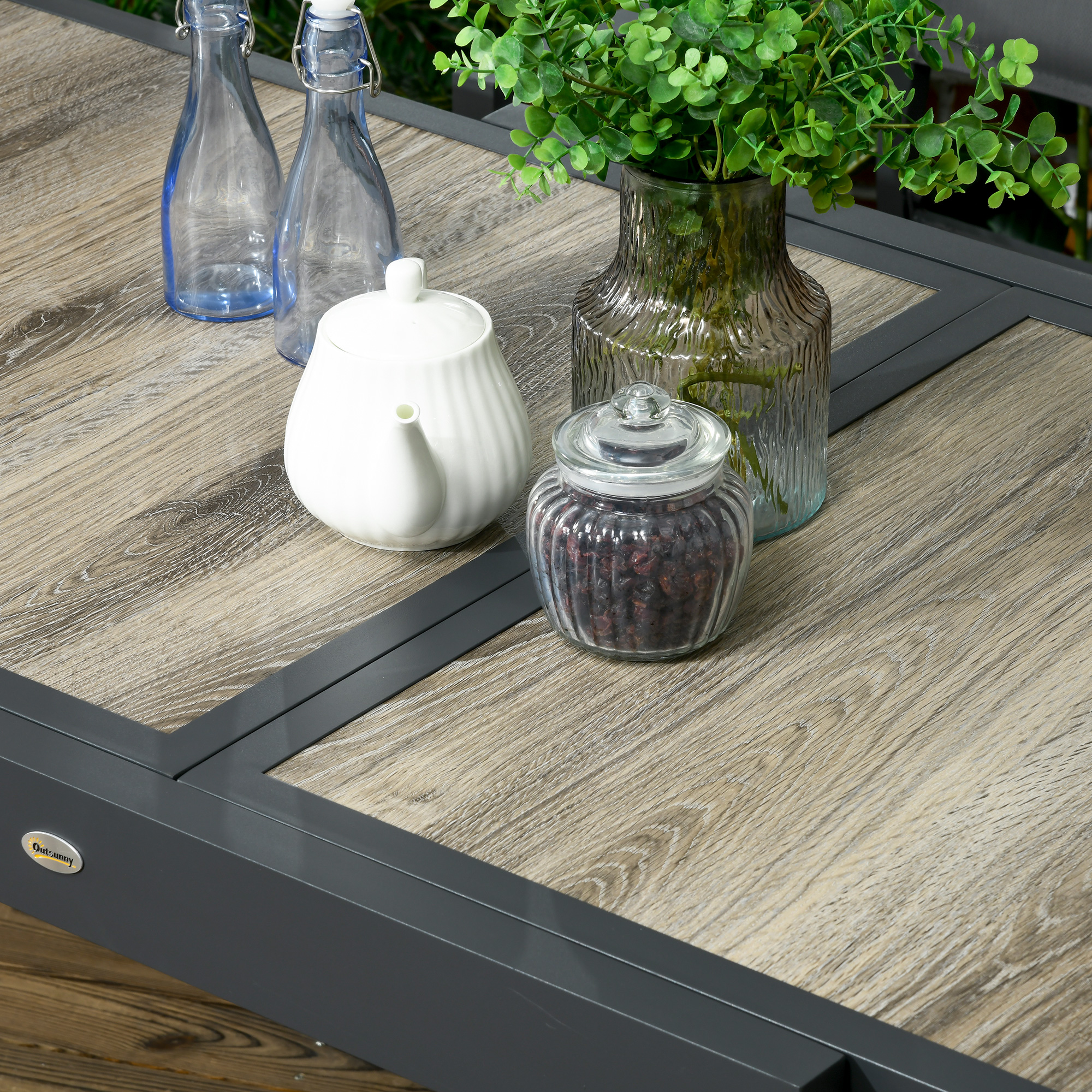Tavolo in legno con due bottiglie di vetro blu, teiera bianca, barattolo di bacche e vaso con piante verdi.