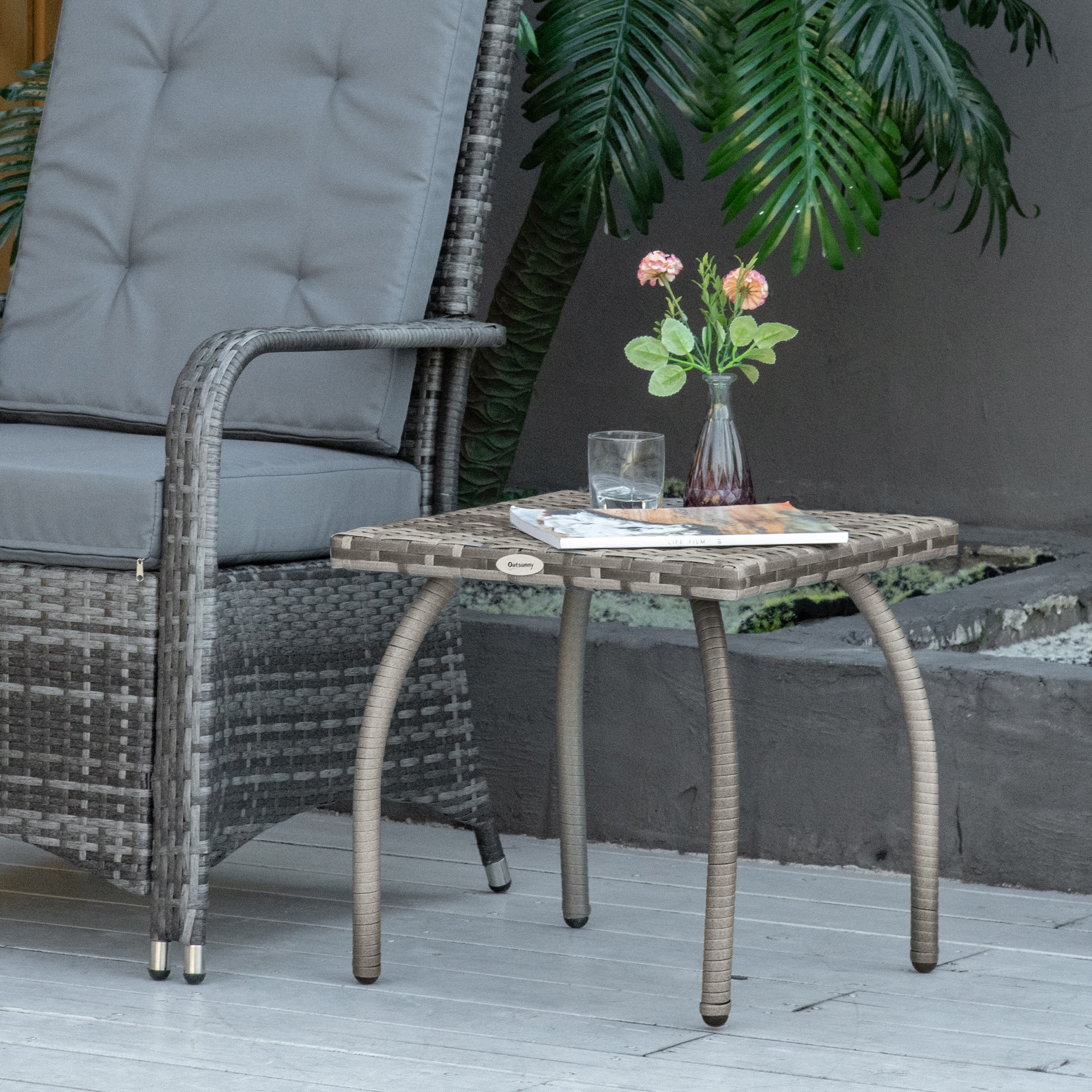 Sedia in rattan grigio accanto a tavolino con vaso, bicchiere e libro, all'aperto.