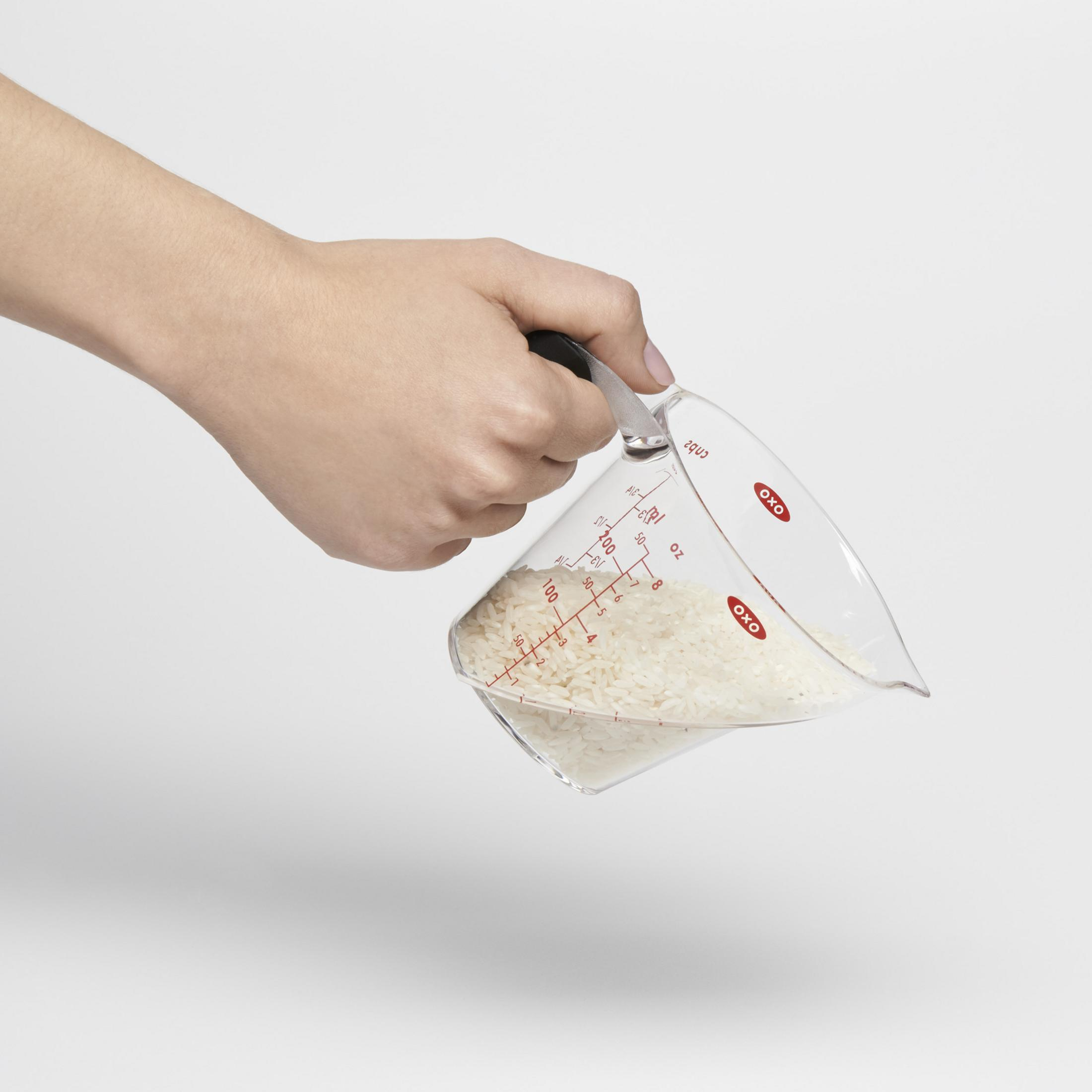 Eine Hand hält einen Messbecher mit Reis, der auf einer weißen Oberfläche steht.