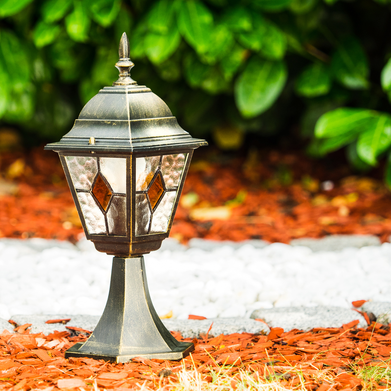 Eine Gartenlampe im Vintage-Stil mit Buntglas steht auf einem Mulchbeet.