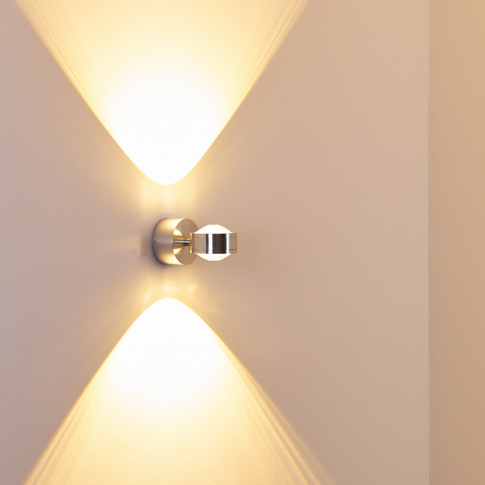 Eine silberne Wandlampe strahlt nach oben und unten Lichtkegel auf eine helle Wand.