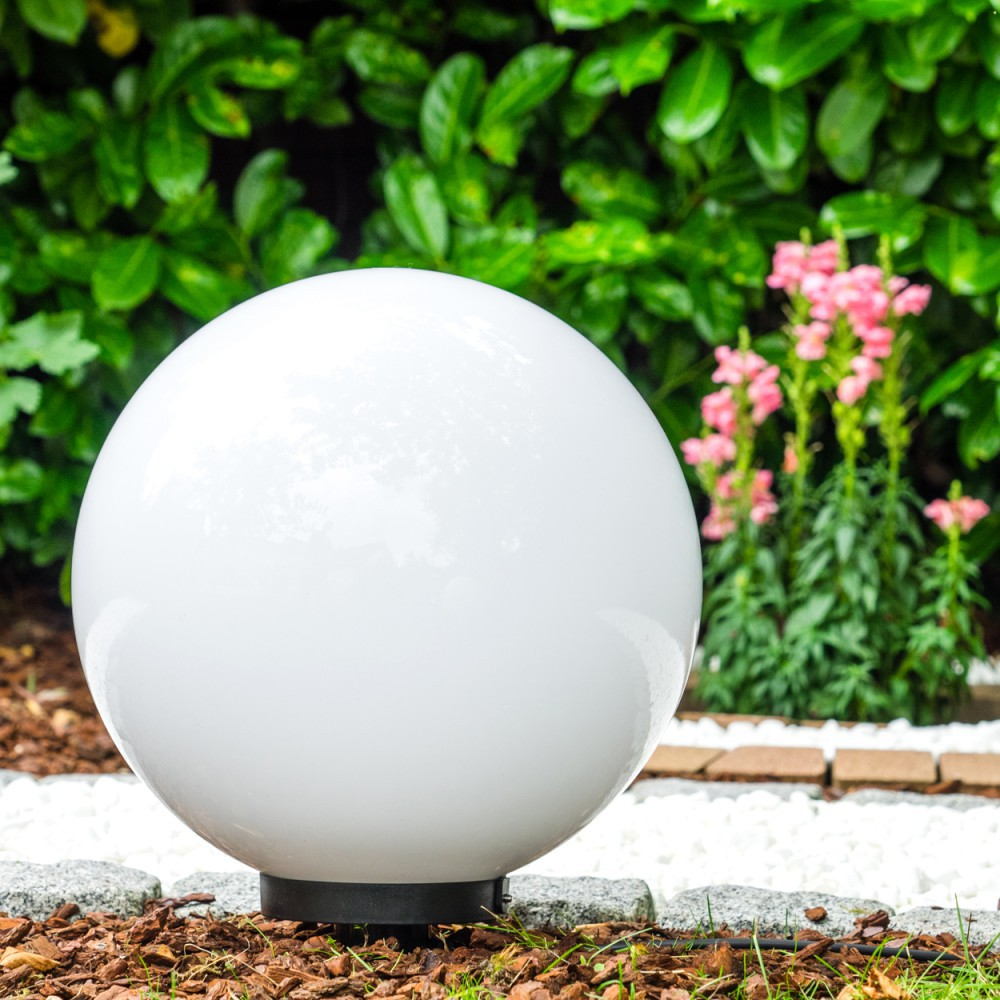 Eine weiße kugelförmige Lampe steht in einem Garten mit grünen Büschen und rosa Blumen.