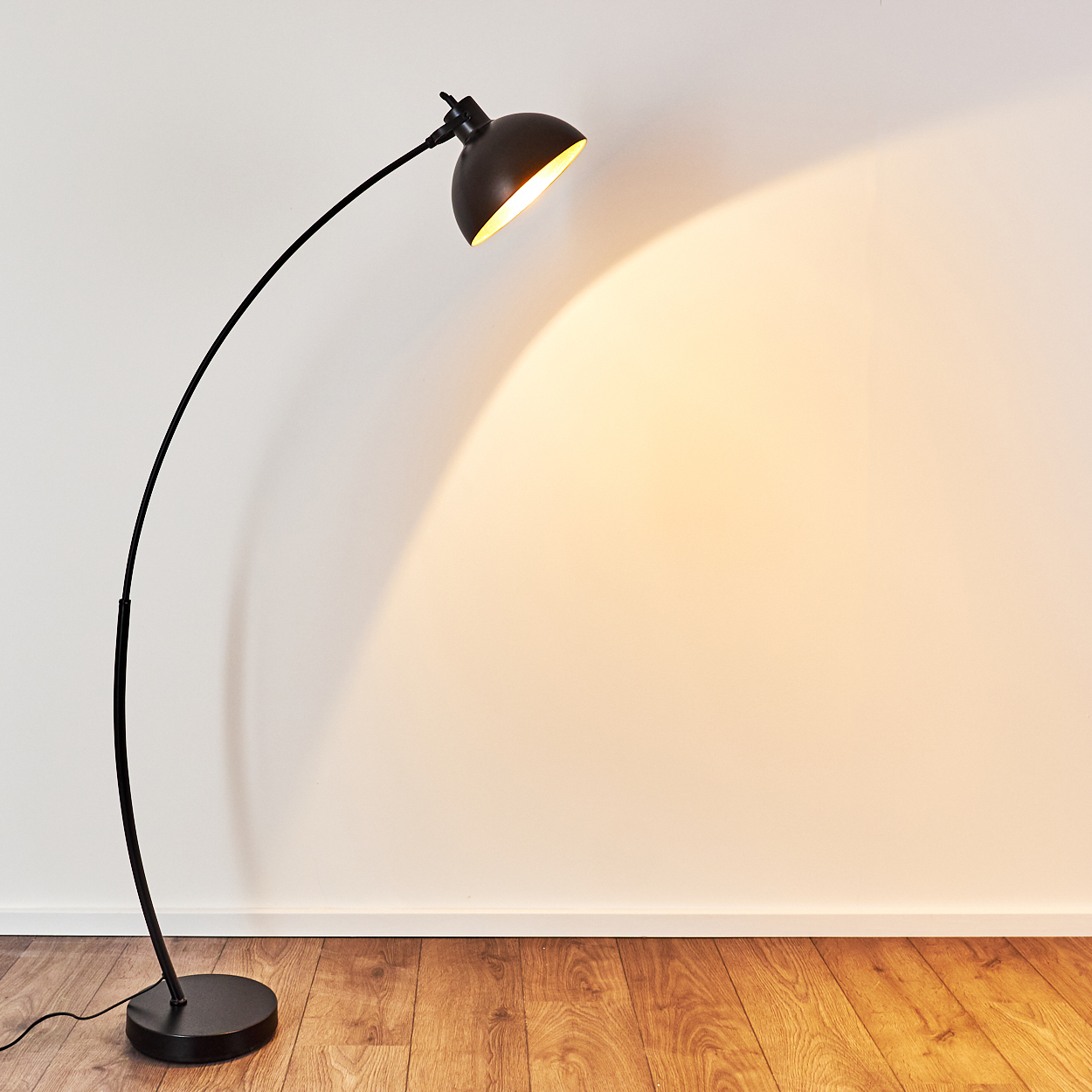Ein schwarzer Bogenlampenfuß steht auf einem Holzboden, die Lampe leuchtet.