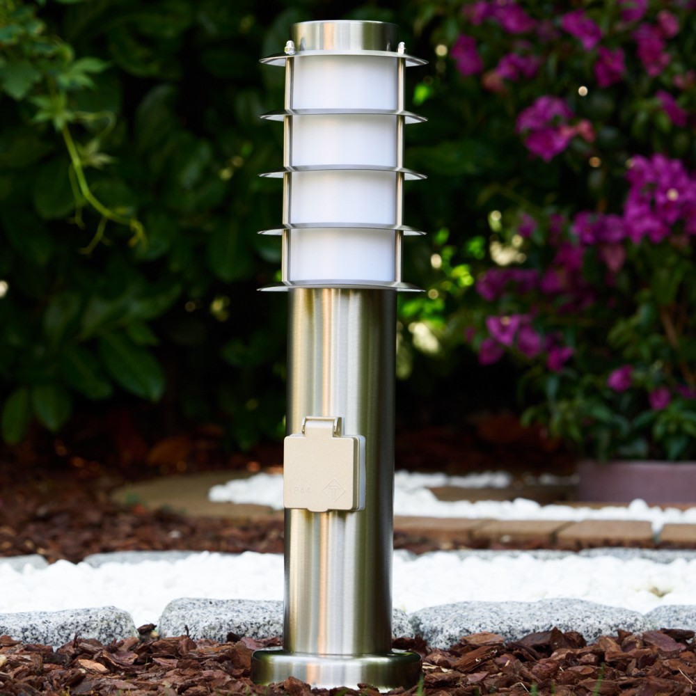 Ein moderner Edelstahl-Gartenlichtmast mit Steckdose, umgeben von dunklem Mulch und Steinen.