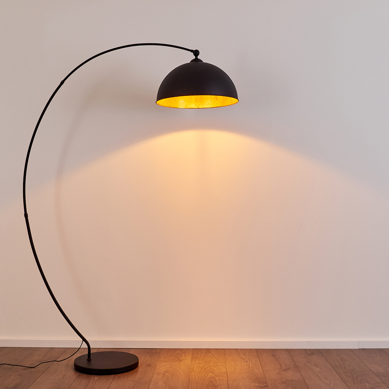 Eine schwarze Bogenlampe mit goldenem Reflektor leuchtet vor einer weißen Wand.