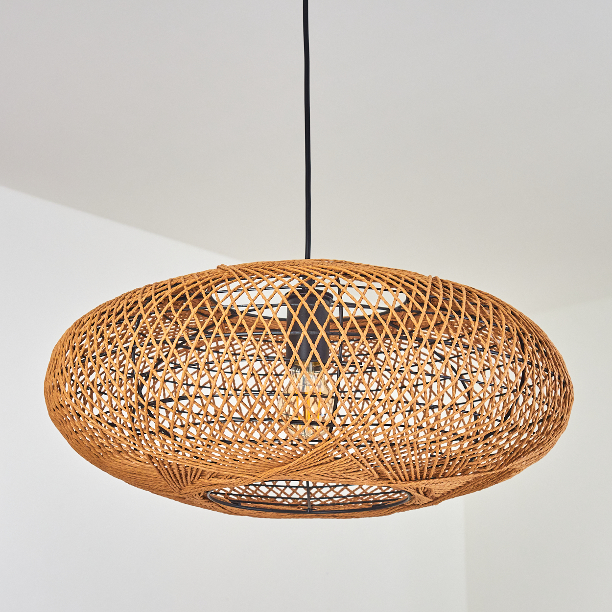 Eine gewebte Rattan-Pendelleuchte mit schwarzem Kabel und sichtbarer Glühbirne hängt drinnen.
