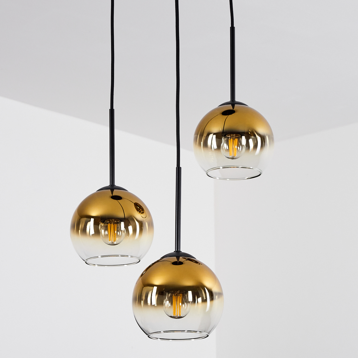 Drei goldene Pendelleuchten mit Glaskugeln und sichtbaren Glühbirnen hängen von schwarzen Kabeln.
