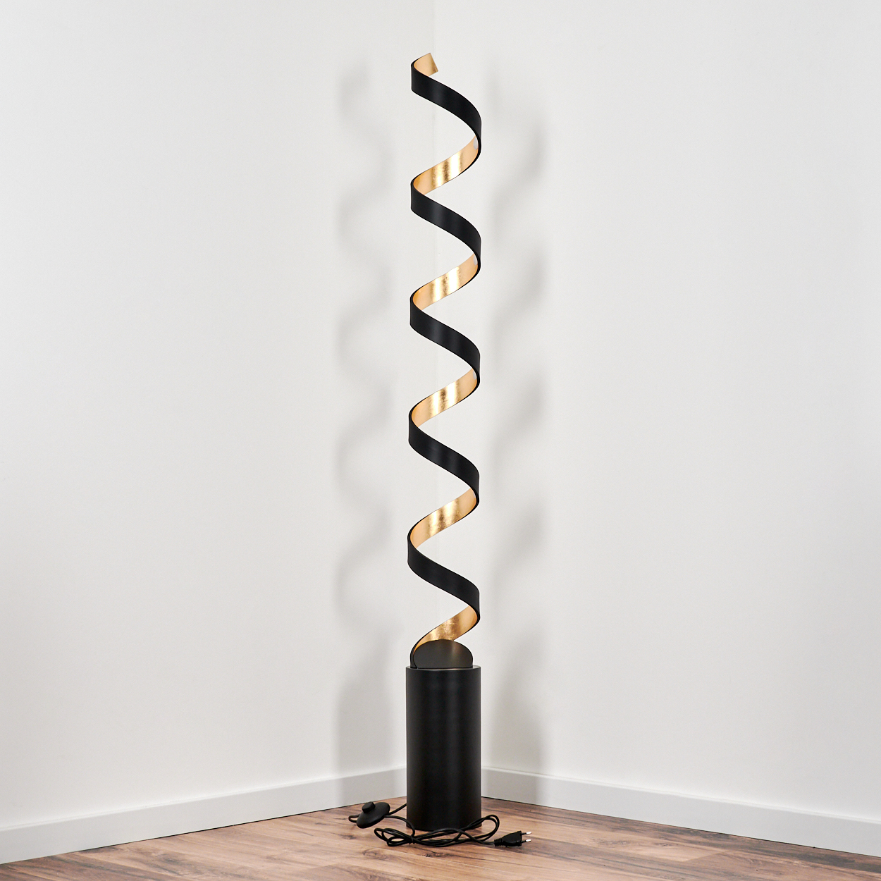 Eine schwarze Stehleuchte mit einem spiralförmigen, gold-schwarzen Band auf einem Holzboden.