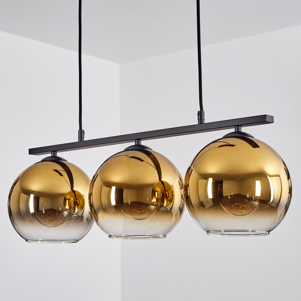 Drei goldene Glas-Pendelleuchten hängen an schwarzen Kabeln an einer schwarzen Stange.
