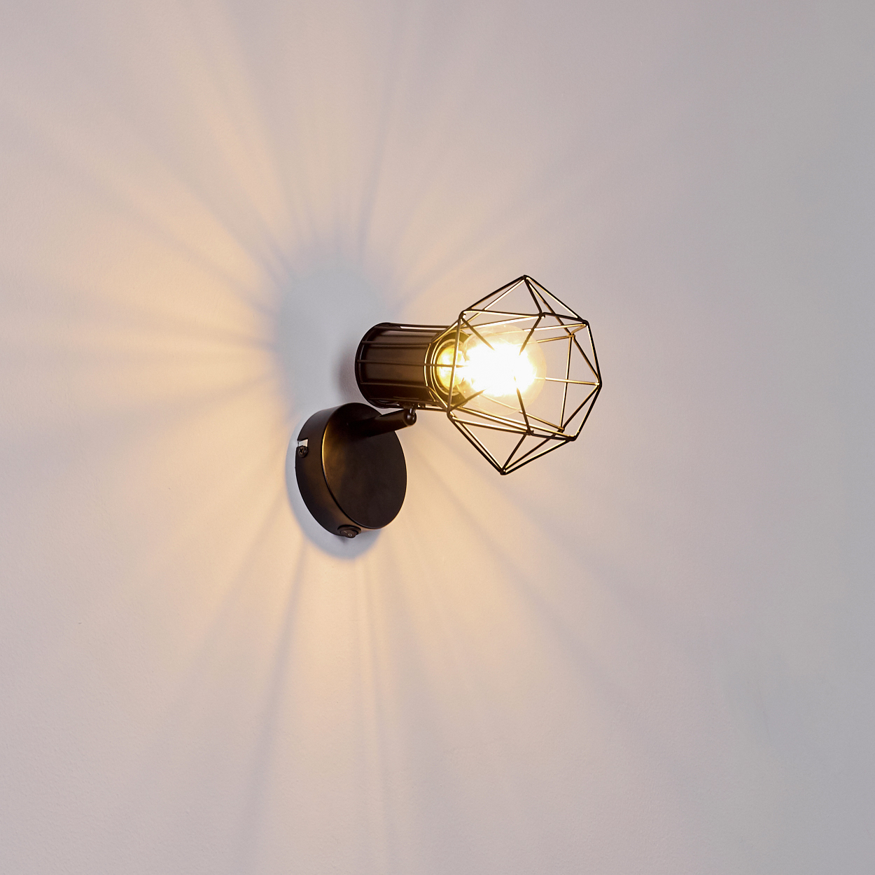 Moderne Wandleuchte mit schwarzem Metallkäfig und Glühbirne, wirft Schatten an die Wand.