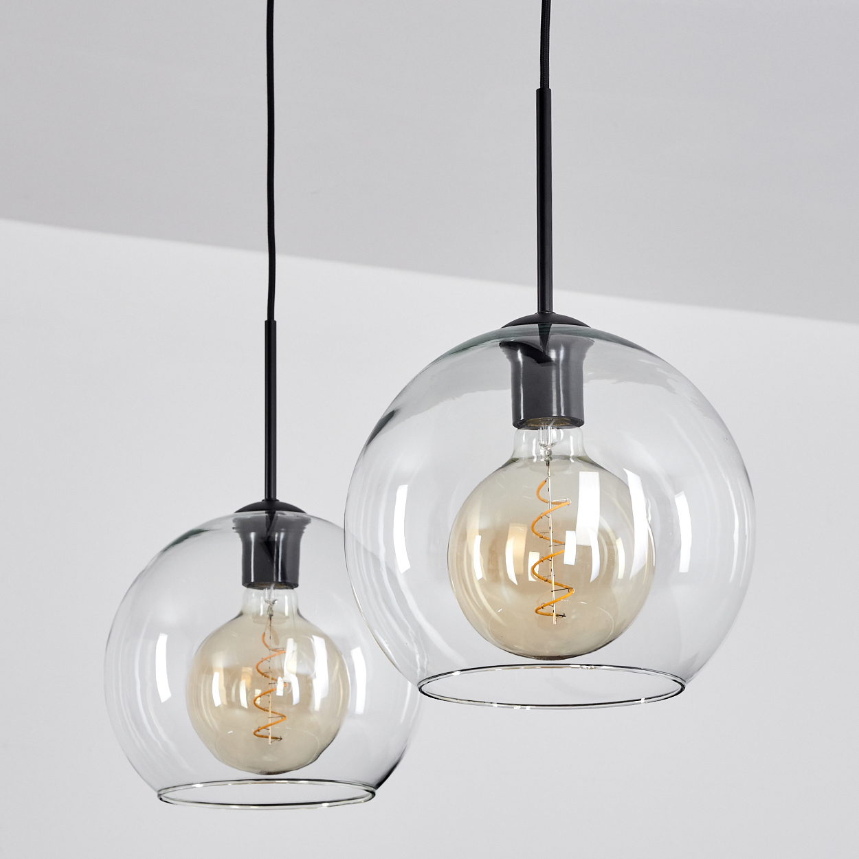 Zwei transparente Glaskugelleuchten mit Edison-Glühbirnen hängen an schwarzen Kabeln.