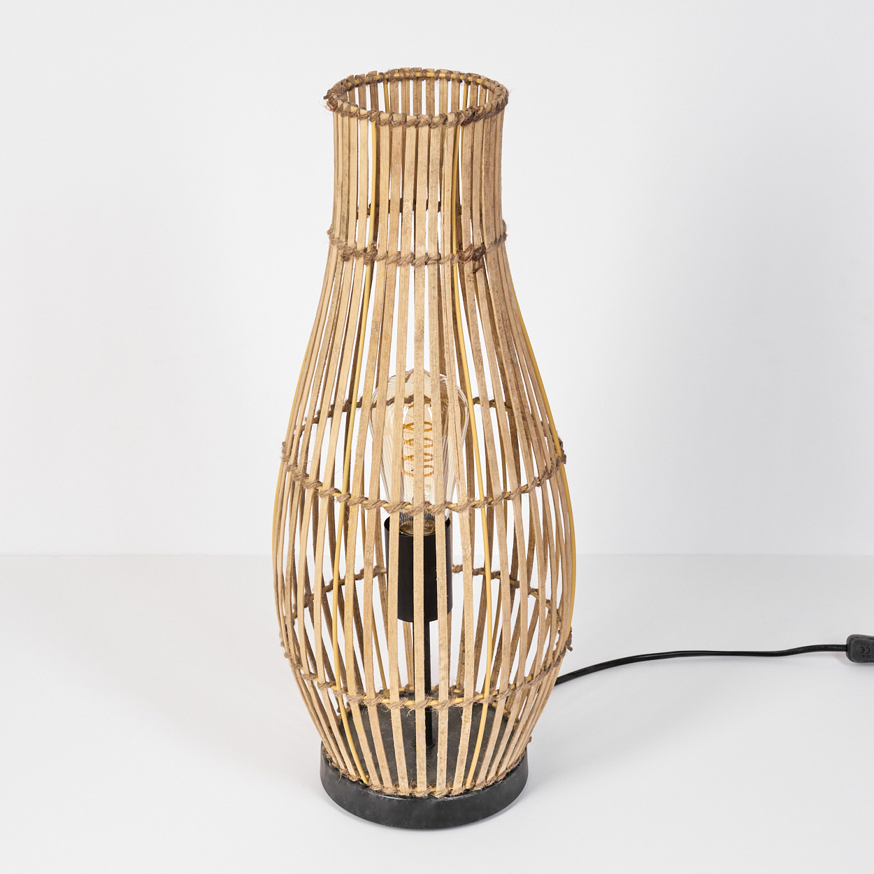 Eine hohe Lampe aus gewebtem Bambus mit sichtbarer Edison-Glühbirne und schwarzem Sockel steht auf weißer Fläche.