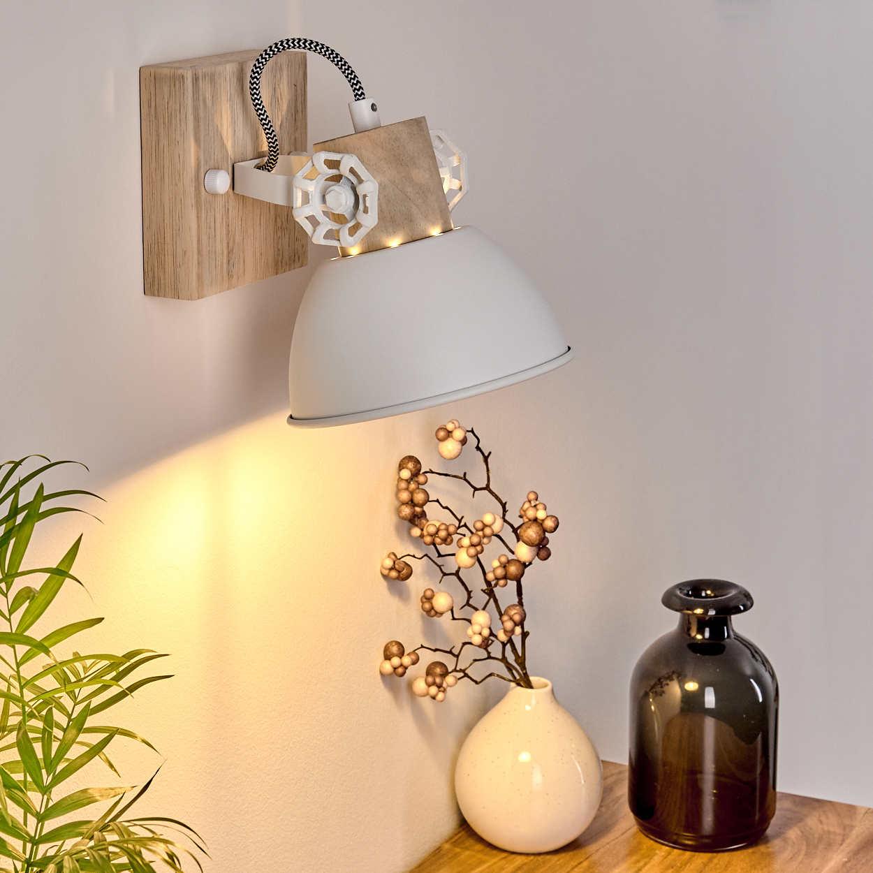 Eine weiße Wandleuchte mit Holzakzenten beleuchtet einen Deko-Zweig in einer Vase.