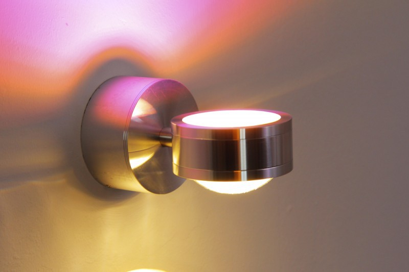Eine moderne Wandleuchte aus Metall wirft farbige Schatten in Pink und Gelb an eine Wand.