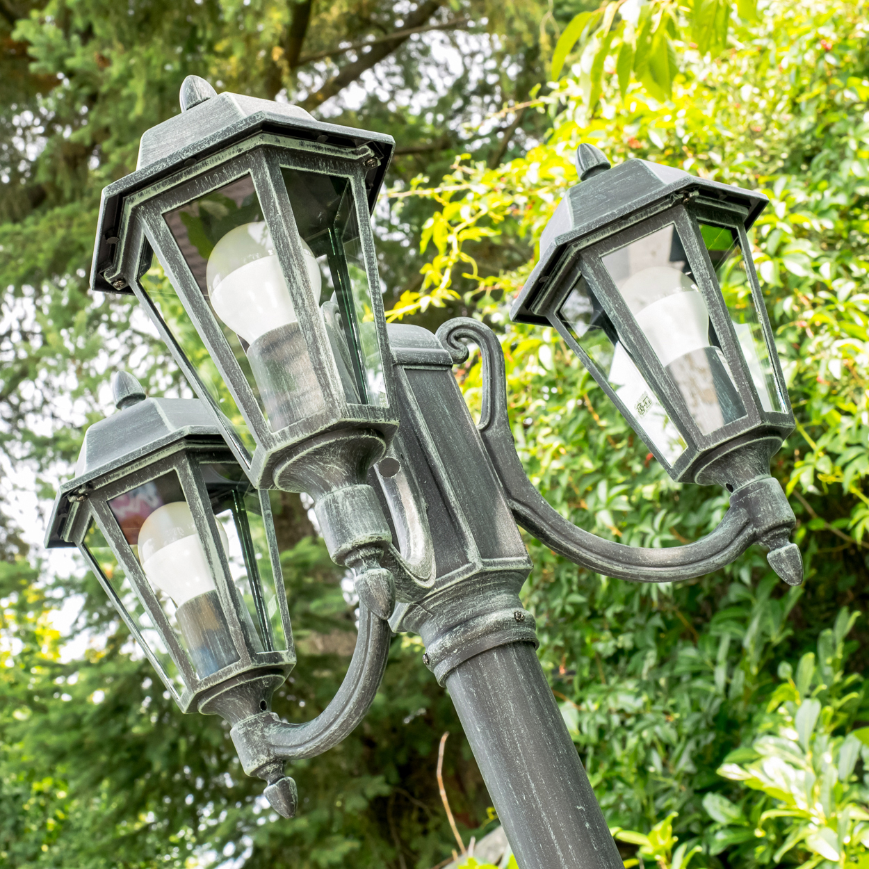 Eine schwarze, dreiköpfige Laterne mit Glaspaneelen und hellen LED-Birnen steht im Freien.