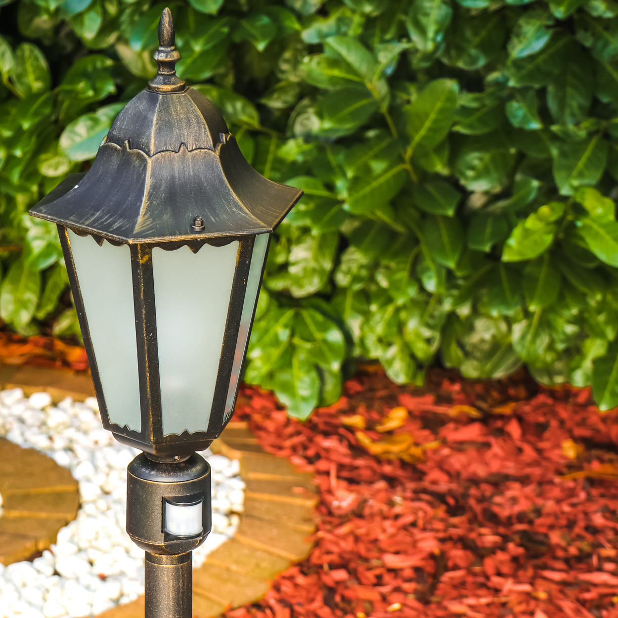 Eine Außenlampe im Vintage-Stil mit Bewegungsmelder steht in einem Garten mit rotem Mulch und weißen Steinen.