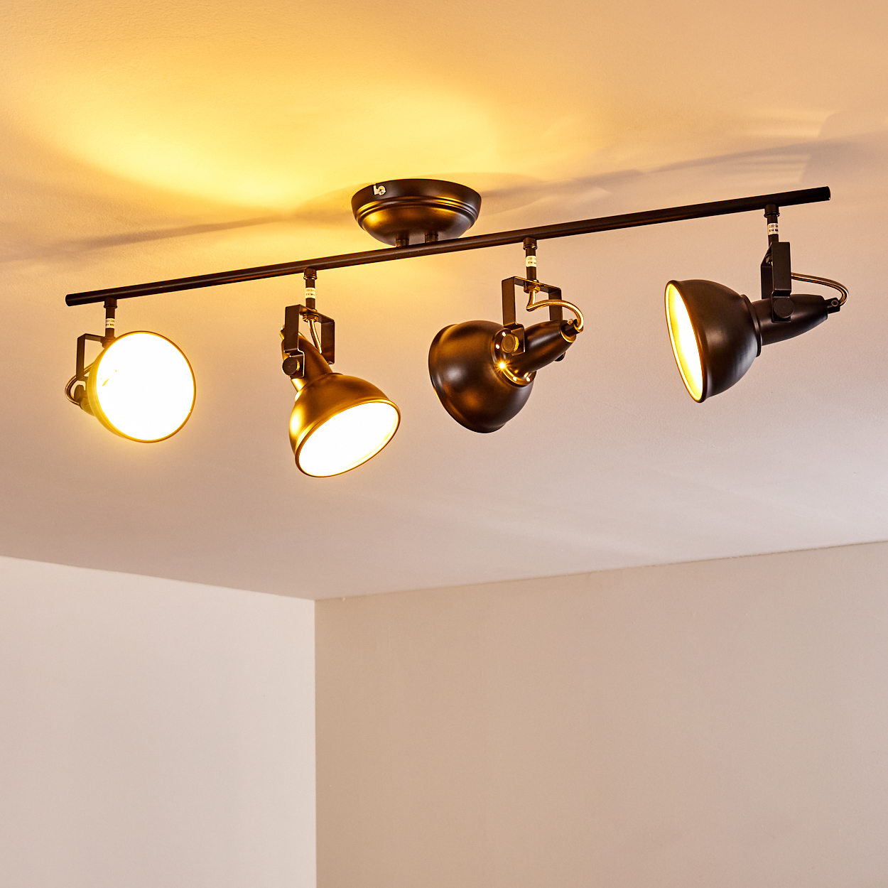Eine schwarze Deckenleuchte mit vier Schienenstrahlern und warmgelben Birnen an einer weißen Decke.