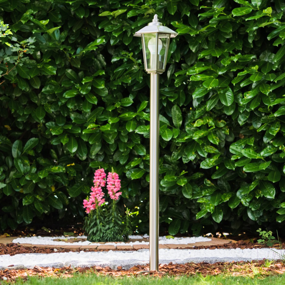 Ein silberner Gartenlaternenpfahl steht vor einer dichten Hecke. Rosa Blumen blühen daneben.