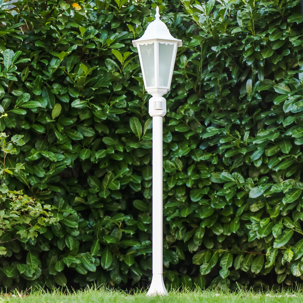 Eine weiße Lampe steht im Gras vor einer grünen Hecke.