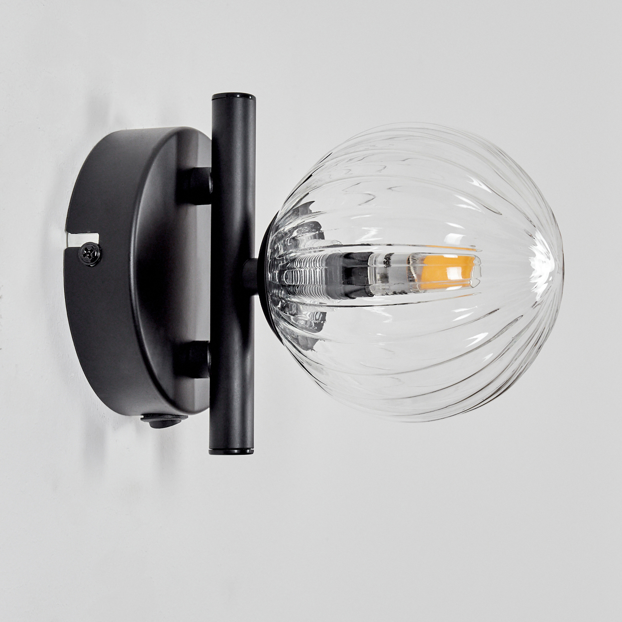 Eine schwarze Wandleuchte mit einem gewellten Glaslampenschirm und einem sichtbaren LED-Leuchtmittel.