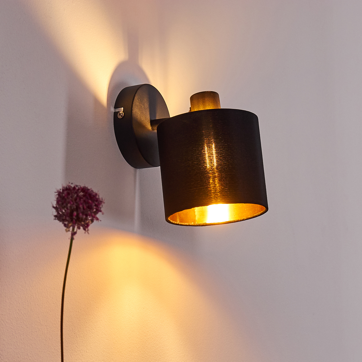 Eine schwarze Wandleuchte mit goldenem Inneren beleuchtet eine weiße Wand. Eine lila Blume ist in der Nähe.