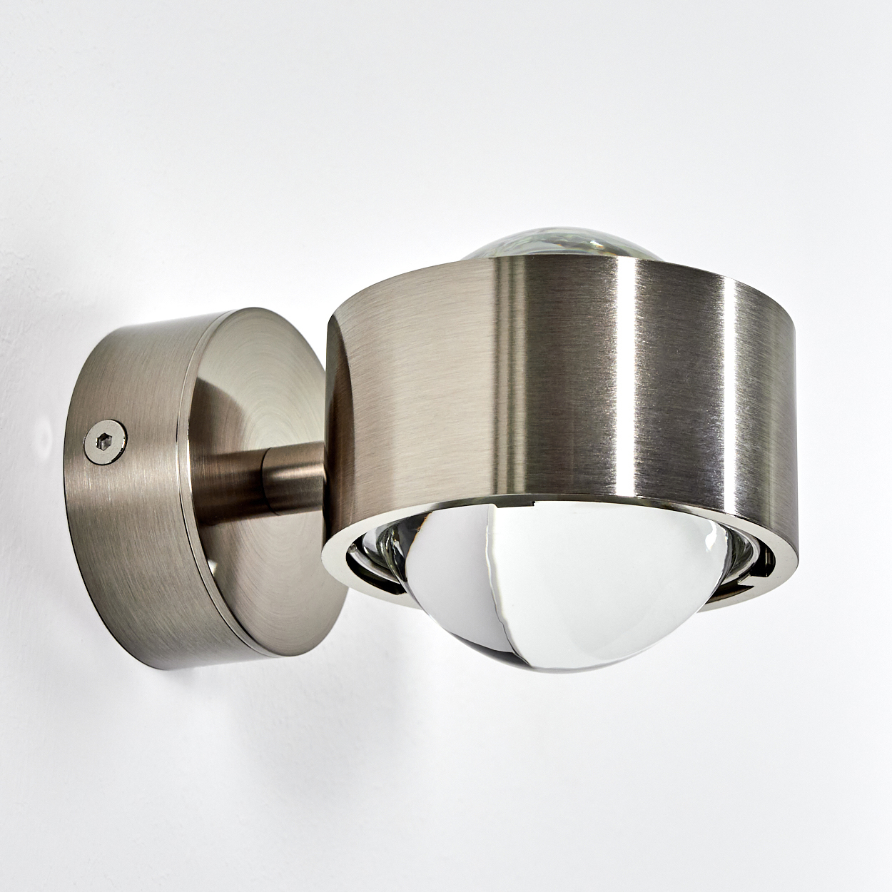 Eine moderne Wandleuchte aus gebürstetem Metall mit einem runden Sockel und einem zylindrischen Lampenkopf.