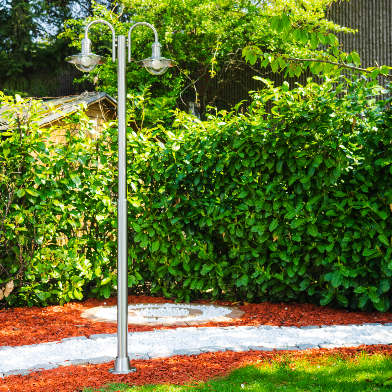Ein zweiflammiger Gartenlichtmast aus Metall steht vor einer dichten Hecke und einem Holzschuppen.