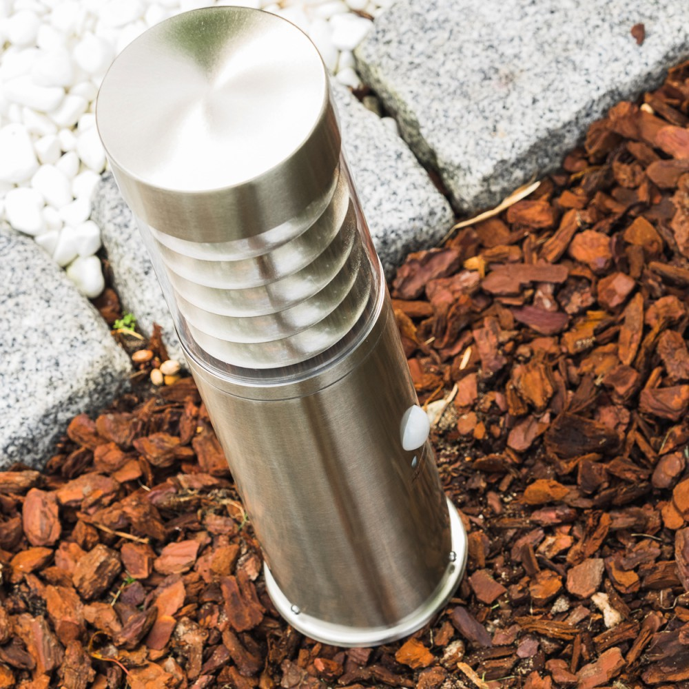 Eine moderne Edelstahl-Gartenleuchte steht auf Rindenmulch, neben Steinen.