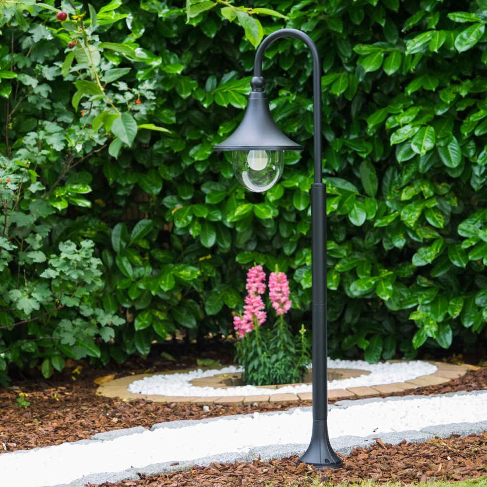 Eine schwarze Gartenlampe steht vor einer üppigen grünen Hecke mit rosa Blumen.