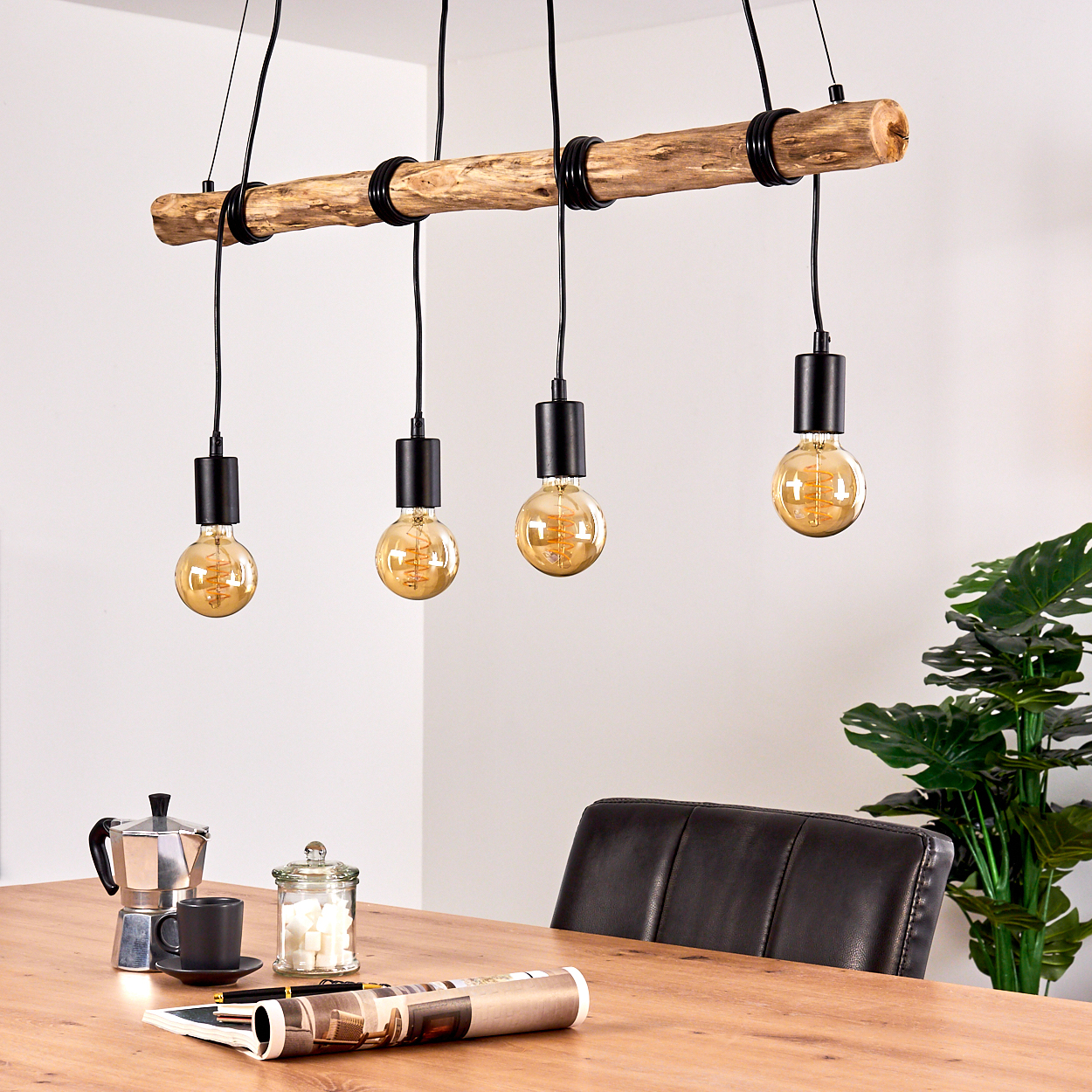 Ein rustikaler Kronleuchter aus Holz mit vier Glühbirnen hängt über einem Holztisch mit Kaffeekanne.