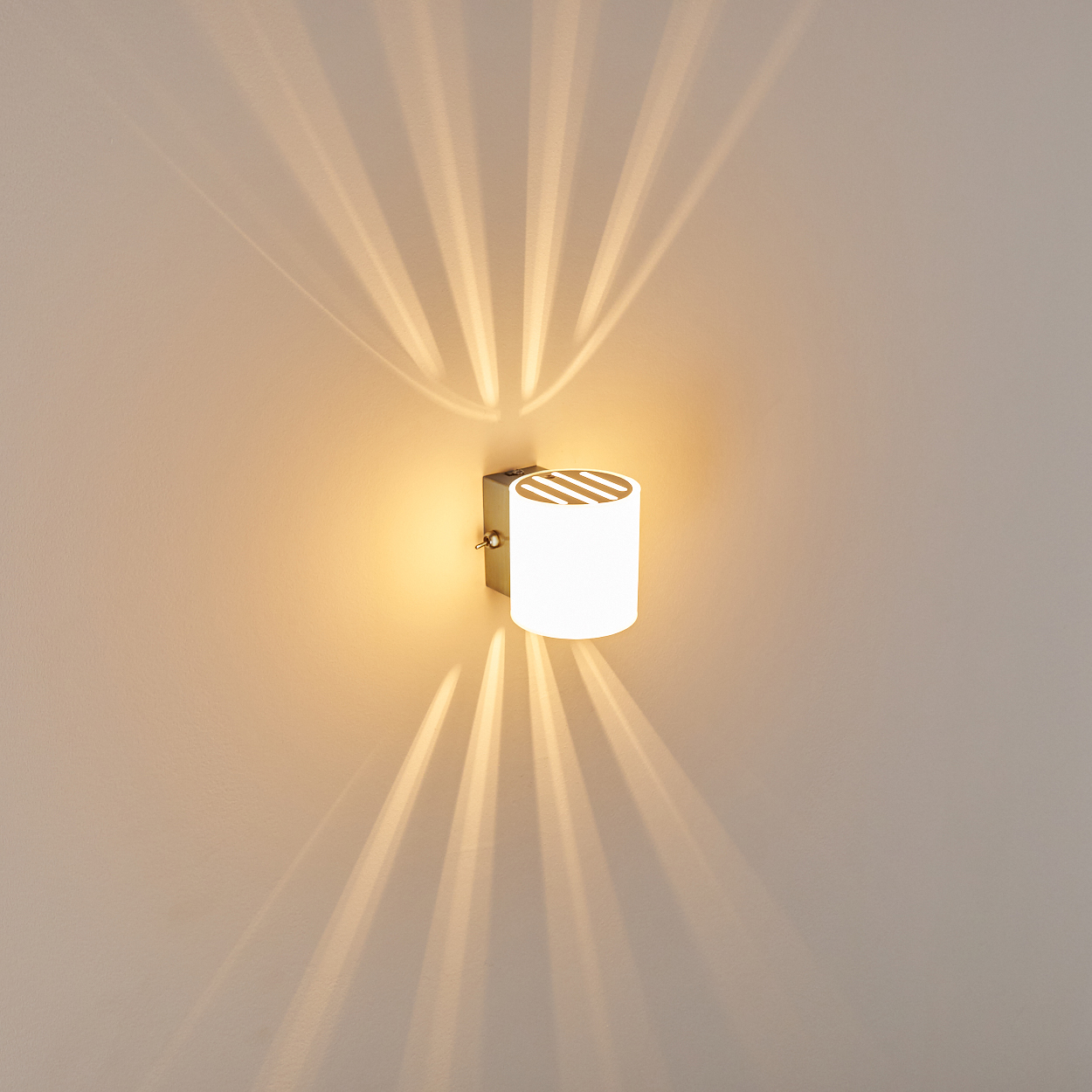 Moderne zylindrische Wandleuchte mit gestreiftem Oberteil, die dramatische Lichtmuster auf eine beige Wand wirft.
