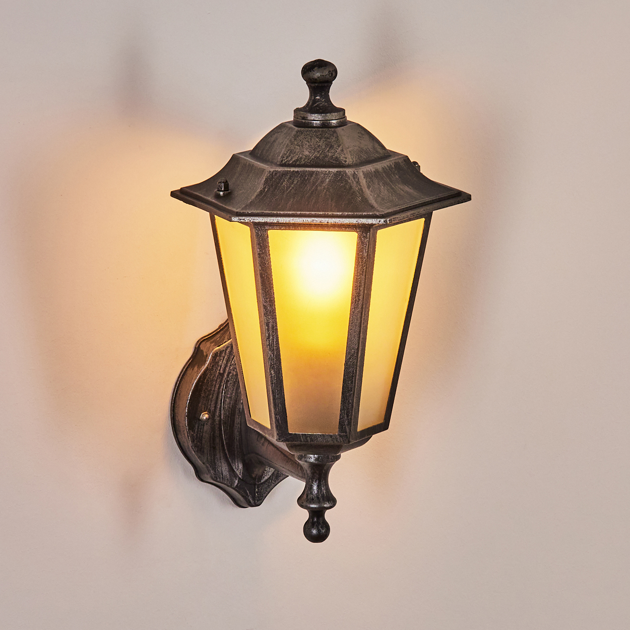 Eine Wandlaterne für den Außenbereich im Vintage-Stil mit dunklem Metallrahmen und Milchglas, beleuchtet von einer warmgelben Glühbirne.