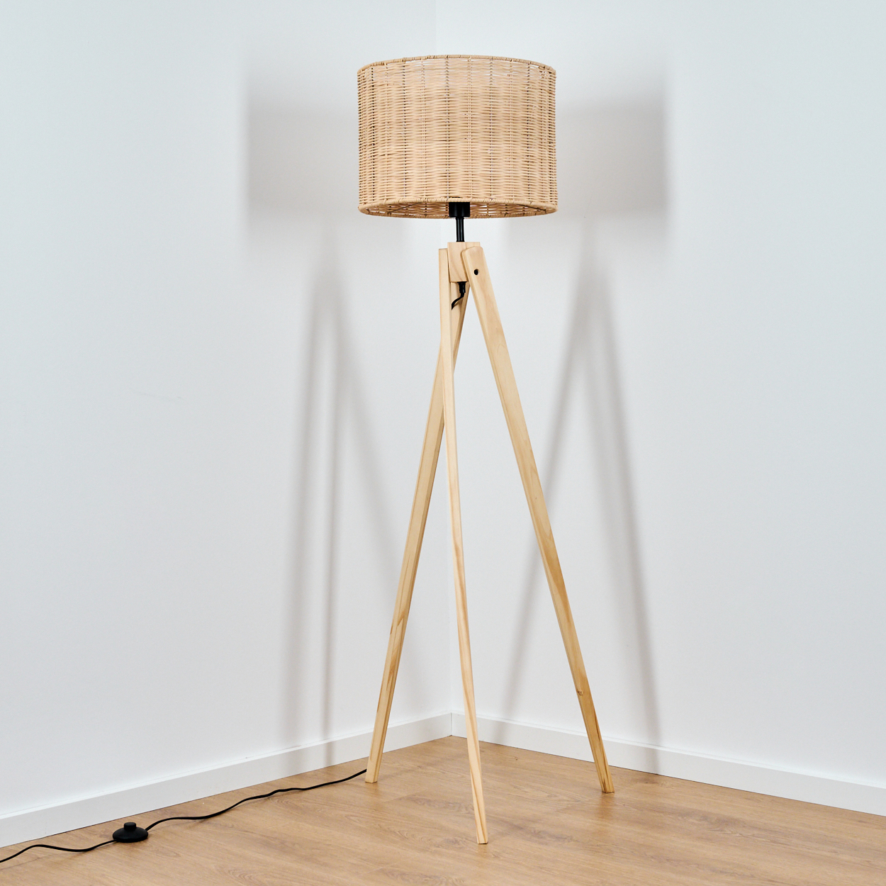 Ein Stehleuchte mit Weidenkorb-Schirm steht auf einem Dreibein aus Holz.