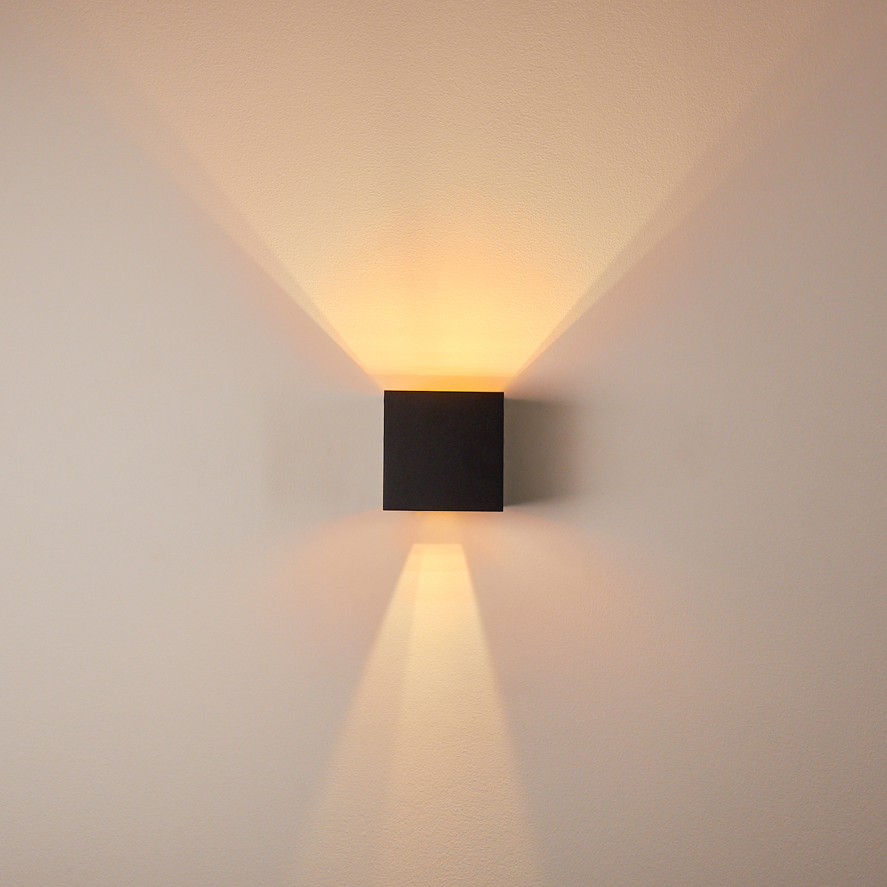 Ein schwarzer kubischer Wandleuchter wirft nach oben und unten warmes Licht an eine weiße Wand.