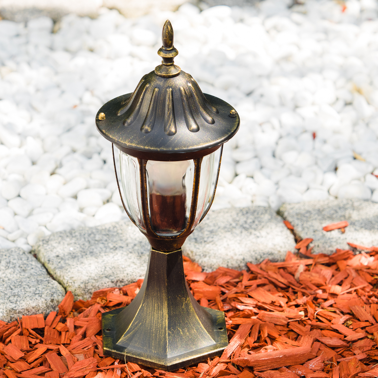 Eine bronzefarbene Lampe steht in roter Rindenmulch und weißem Kies.