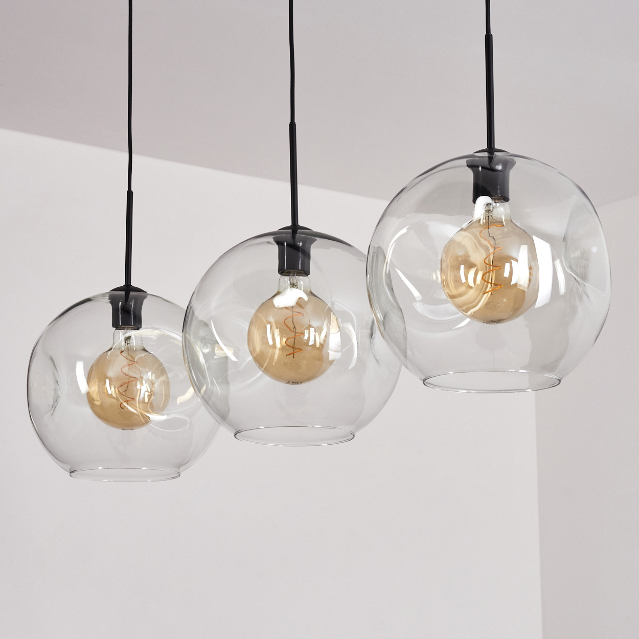 Drei transparente Glaskugelleuchten mit Edison-Glühbirnen hängen an schwarzen Kabeln.