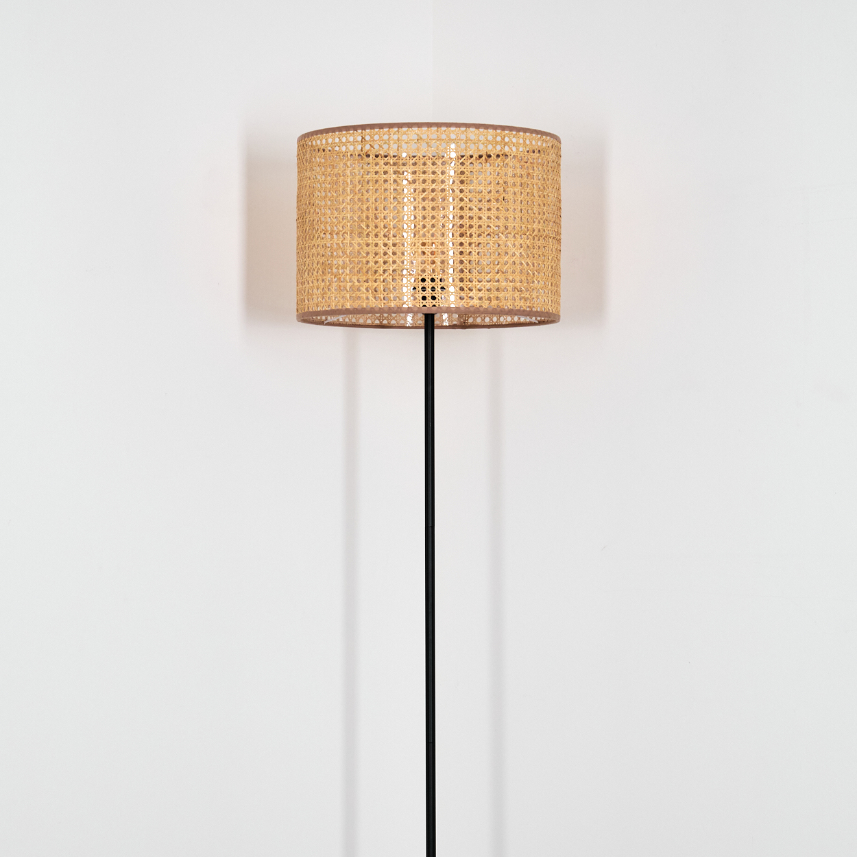 Eine Stehlampe mit einem Korbflecht-Schirm und einem schlanken schwarzen Ständer steht vor einer weißen Wand.