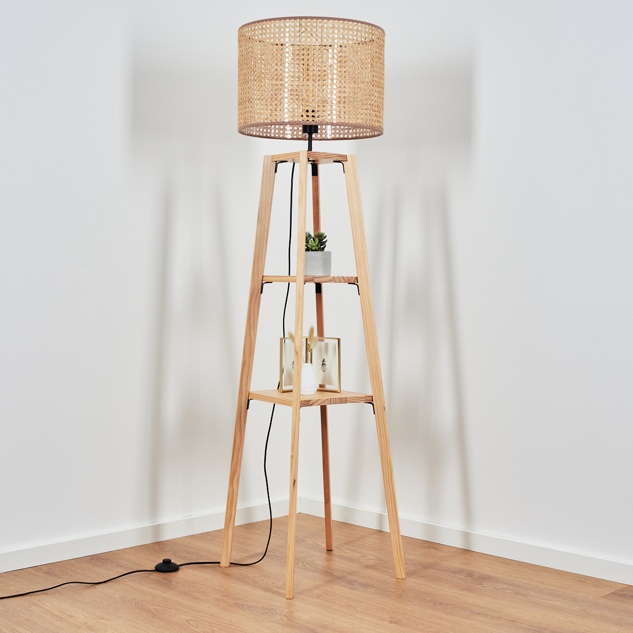 Ein Holz-Stehlampe mit Rattangehäuse, zwei Regalen mit Deko und einem Fußschalter.