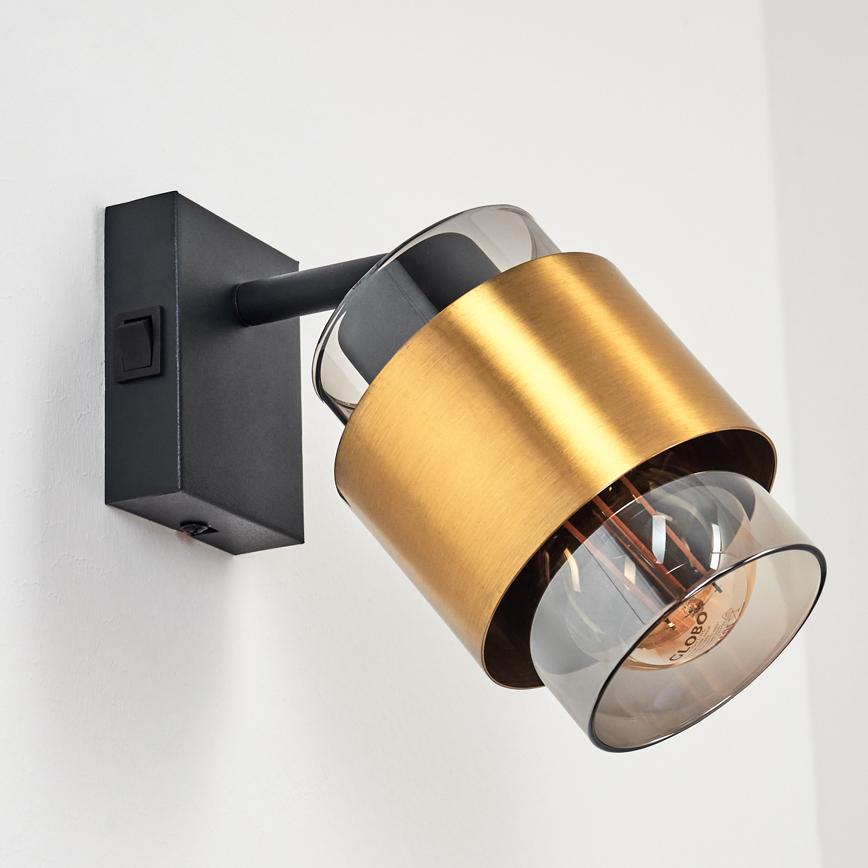 Moderne Wandleuchte mit schwarzem Sockel, goldfarbenem Zylinder und rauchglasigem Lampenschirm.